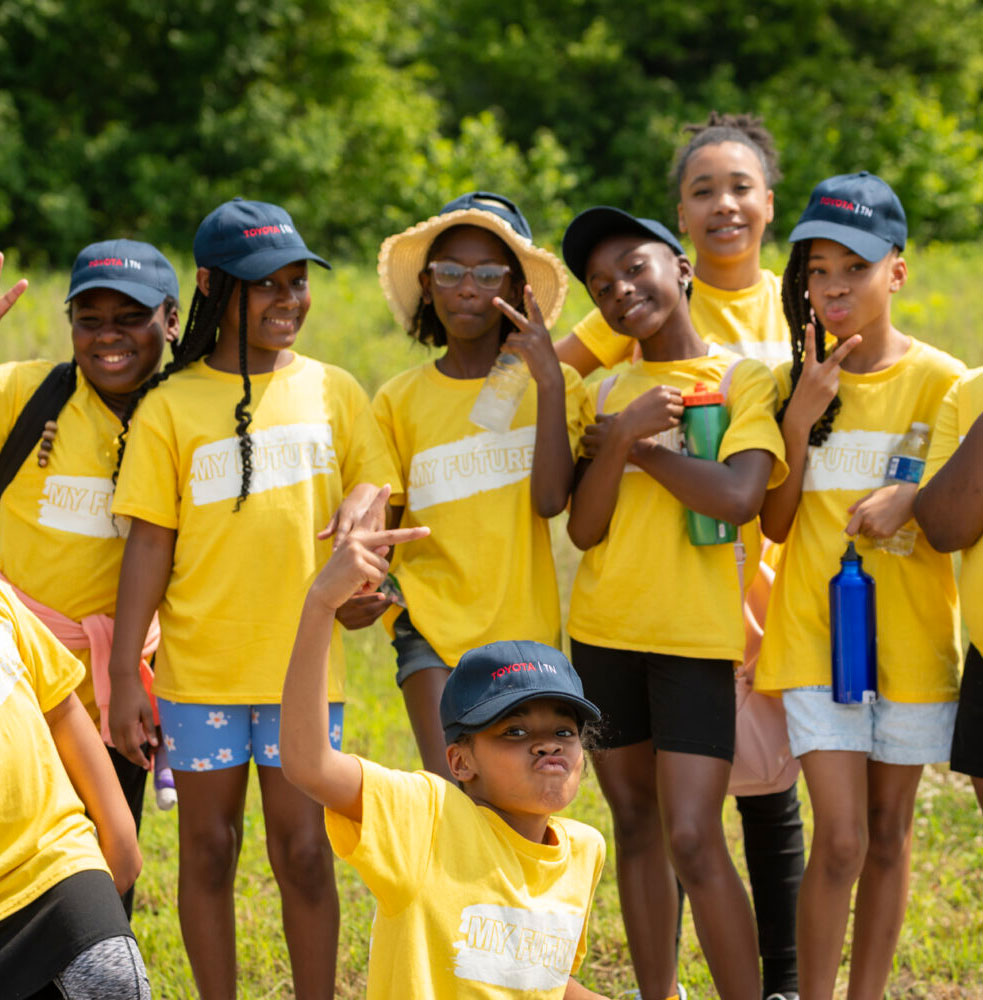 Photo of seven kids posing and smiling in a sunny field, wearing Toyota hats and wearing yellow shirts that read "My Future"