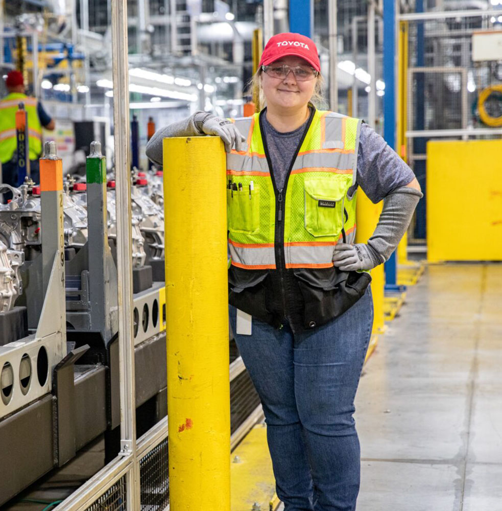 Photo of a Toyota West Virginia worker posing next to an assembly line. She is leaning on a yellow bollard and is wearing a red Toyota hat and PPE